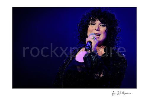 Horizontal color image of Ann Wilson singing into a microphone during a Heart concert in a close-up portrait under deep blue stage light