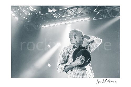 Horizontal black and white image of Gord Downie of The Tragically Hip in an emotional live performance moment, holding his hat to his chest with one hand behind his neck under soft stage light