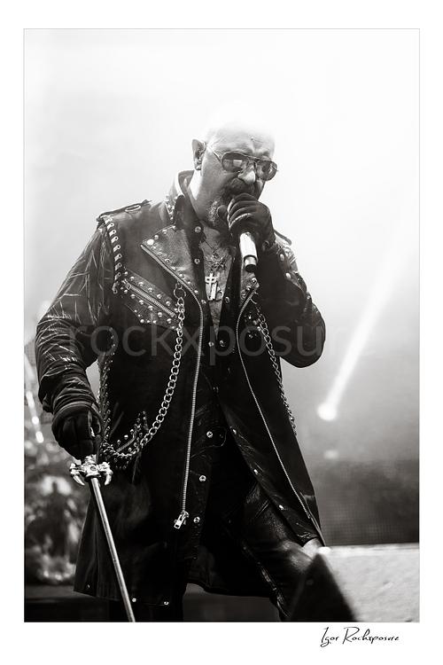 Vertical black and white image of Rob Halford of Judas Priest performing live on stage, singing into a microphone while wearing a studded leather coat and sunglasses