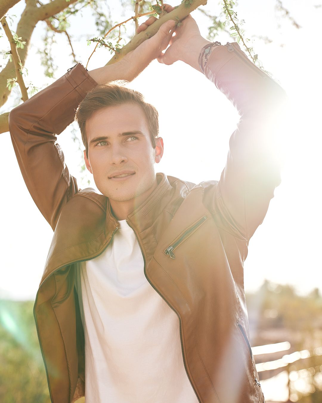 Male model wearing a camel colored leather jacket with his hands above his head grasping a tree branch and looking off camera with the sun setting and providing a solar flair behind his left elbow