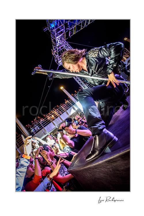 Vertical color image of Joey Tempest of Europe performing live on stage in a black leather jacket with a microphone stand, low-angle perspective, and photographers gathered below