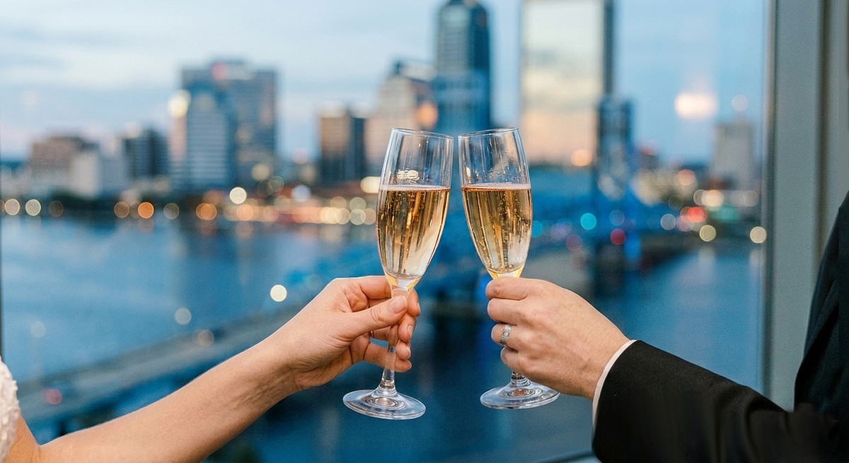 Fine art wedding photography detail of two crystal champagne flutes toasting against a scenic modern city skyline at sunset, with soft blue hour lighting.