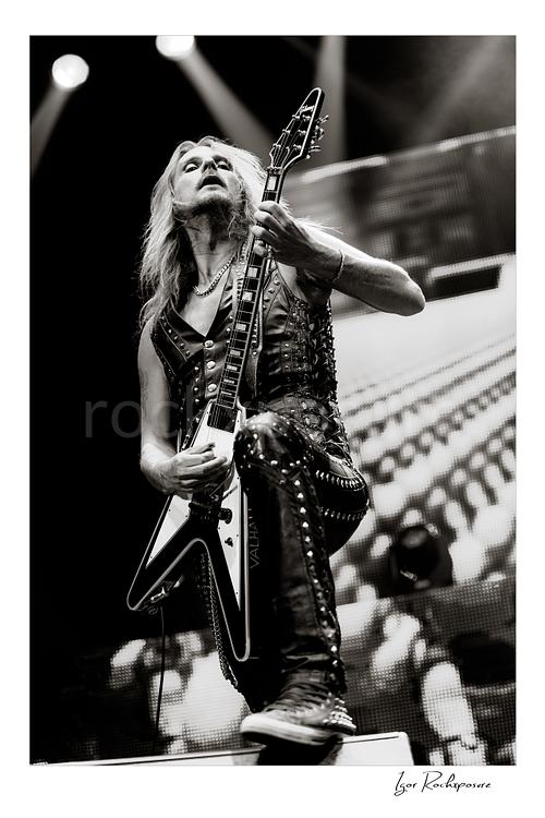 Vertical black and white image of Richie Faulkner of Judas Priest performing live on stage, playing flying V guitar in a dramatic low-angle shot under bright concert lighting