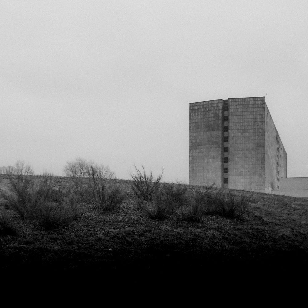 Czarno-biała fotografia autorstwa Michała Sumki ukazująca minimalistyczny krajobraz na Polach Mokotowskich w Warszawie. W tle widoczny surowy, geometryczny budynek na tle pustego, pochmurnego nieba.