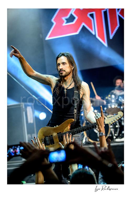 Vertical color image of Nuno Bettencourt performing live on stage with an electric guitar, pointing outward above the crowd
