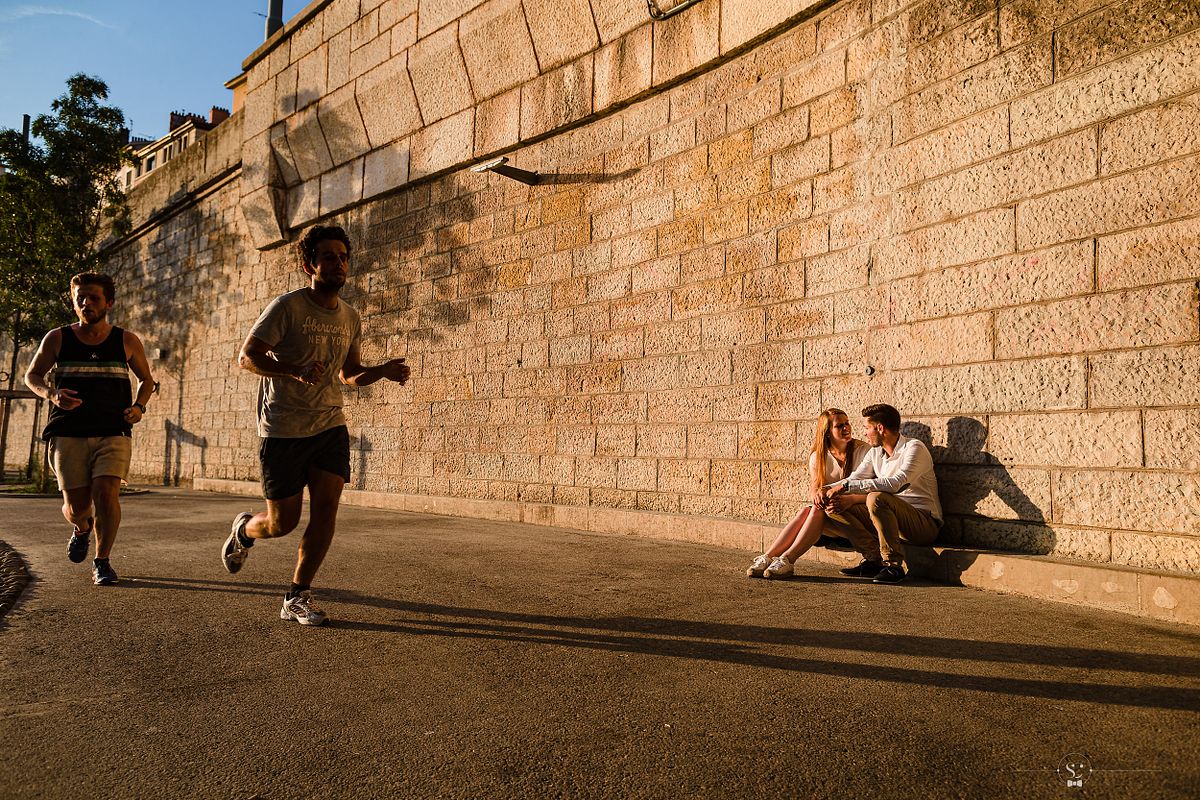 Tarif Photographe Mariage - Sebastien CLAVEL Photographe - Couple assis contre un mur de pierres pendant que deux hommes courent, capturant un moment de complicit&eacute; en ville