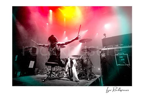 Horizontal color image of Zoltan Chaney at the drum kit with arms outstretched under red, yellow, and white stage lights