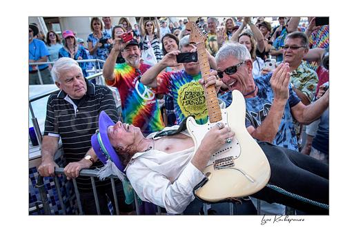 Horizontal color image of Randy Hansen playing a white guitar while leaning back into the crowd during an outdoor ship deck performance