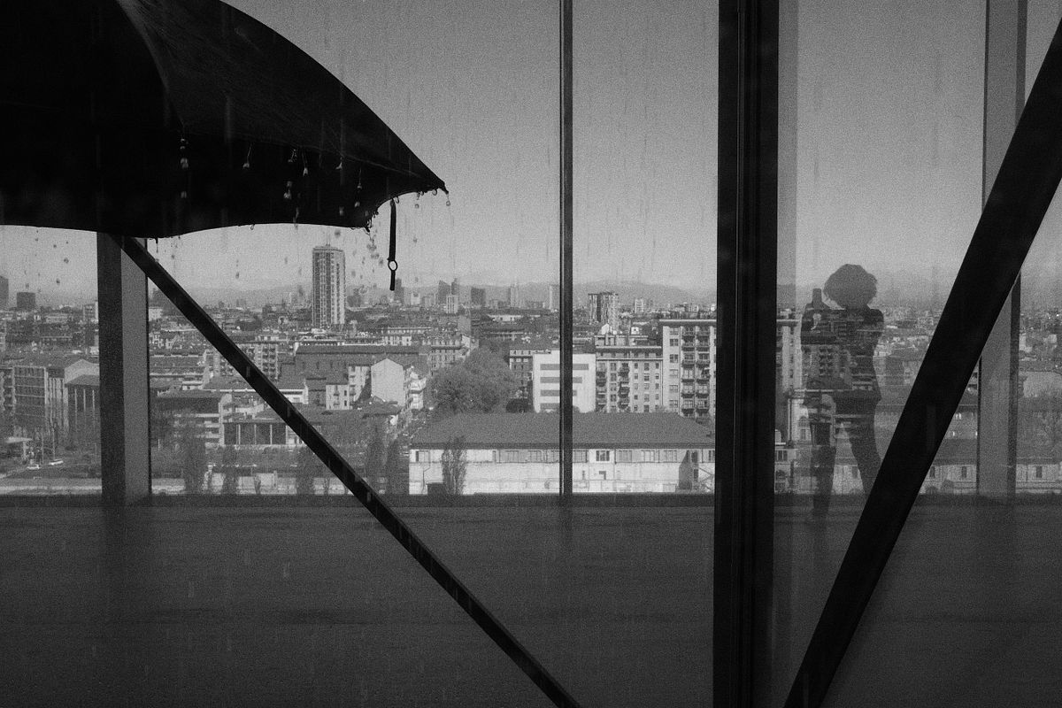 A reflection of a person with an umbrella in a window, showing a cityscape, captured indoors by photographer Sandeep Gajula