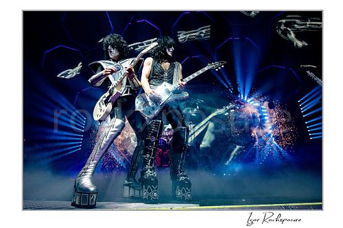 Horizontal color photograph of Paul Stanley and Tommy Thayer performing side by side in full makeup and costumes with bright stage lighting, hexagonal screens, and pyrotechnics behind them