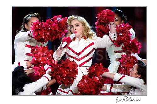 Horizontal color image of Madonna performing with dancers and bright red pom-poms in a theatrical live stage scene