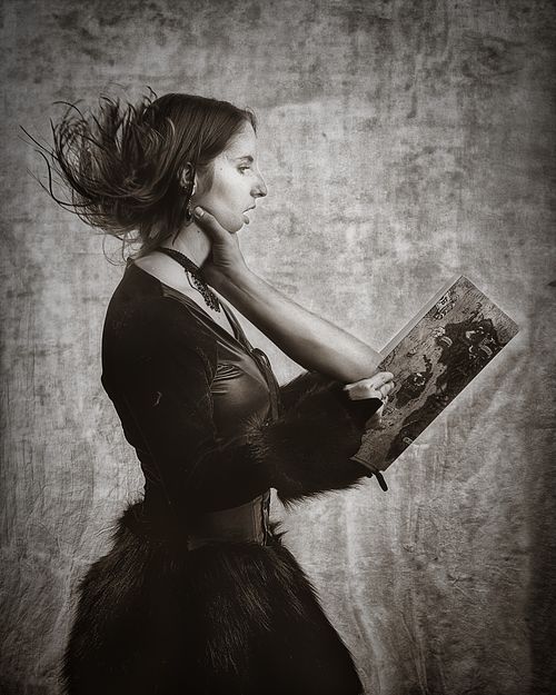 Portrait artistique en noir et blanc d’une femme en costume tenant un livre dont une main semble émerger, la saisissant à la gorge, évoquant une absorption surréaliste par la fiction — photographie fine art par Pascal Yennis, Namur, Belgique.