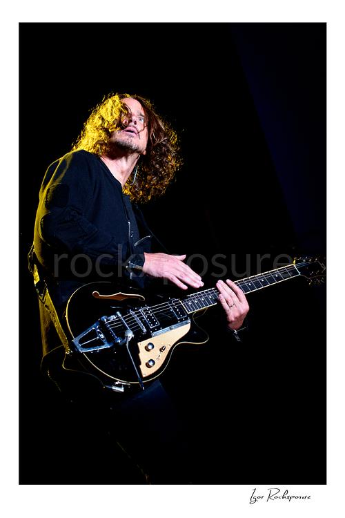Vertical color image of Chris Cornell performing live on stage, playing electric guitar against a dark background under warm concert lighting