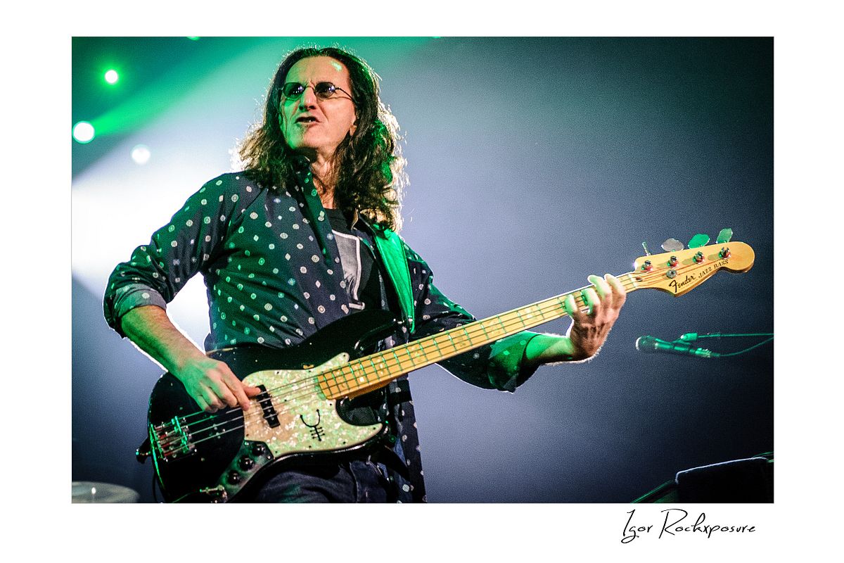 Horizontal concert photography of Geddy Lee playing bass guitar under green stage lighting