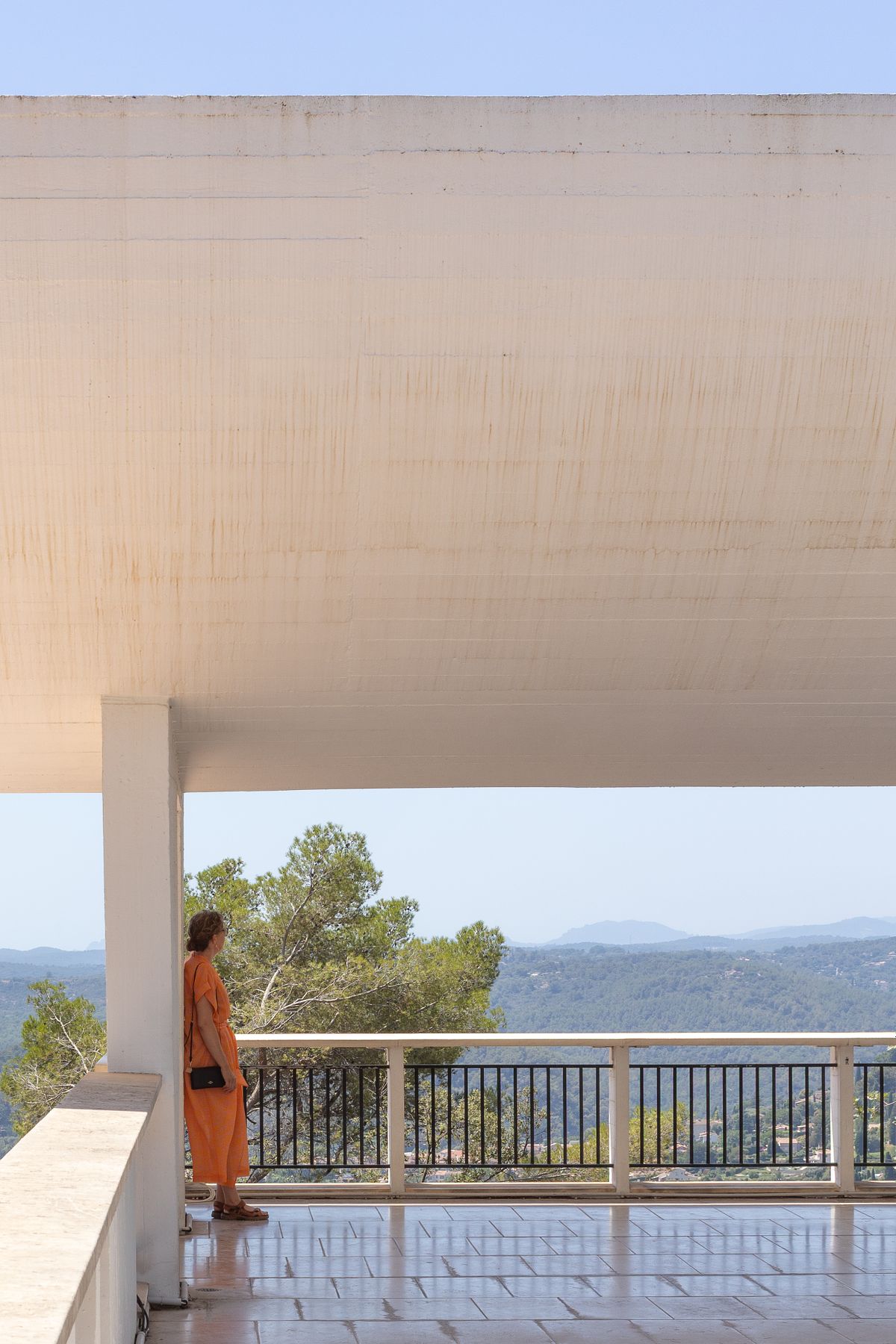 Kvinna i orange kl&auml;nning st&aring;r under ett stort tak och blickar ut &ouml;ver berg och dalg&aring;ng.