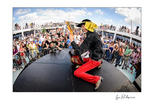 Horizontal color image of Randy Hansen kneeling on an outdoor ship deck stage with guitar and a large crowd behind him