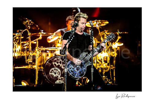 Horizontal color image of Chad Kroeger of Nickelback playing guitar onstage with the drummer visible behind him under warm concert lighting