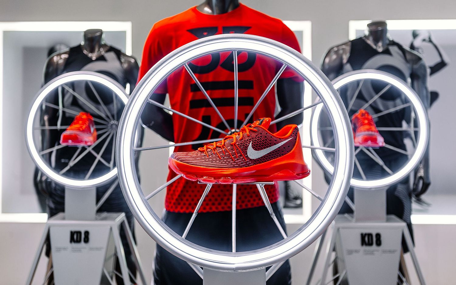 Commercial retail display photograph of the Nike KD 8 'Hunt's Hill Sunrise' sneaker suspended in a white iluminated circular rim, set against a backdrop of mannequins in Nike athletic apparel