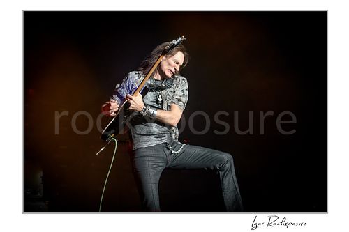 Horizontal color image of Steve Vai playing guitar with one leg raised in a dramatic stage pose under warm concert lighting