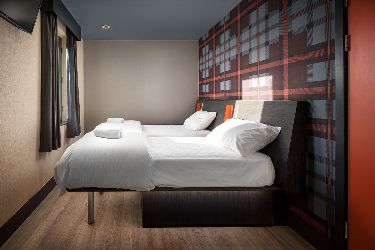 Interior view of a twin room at the Easy Hotel Dublin. Two beds with white linens and dark headboards are positioned against a wall with a red and grey geometric pattern. Light wood-effect flooring.