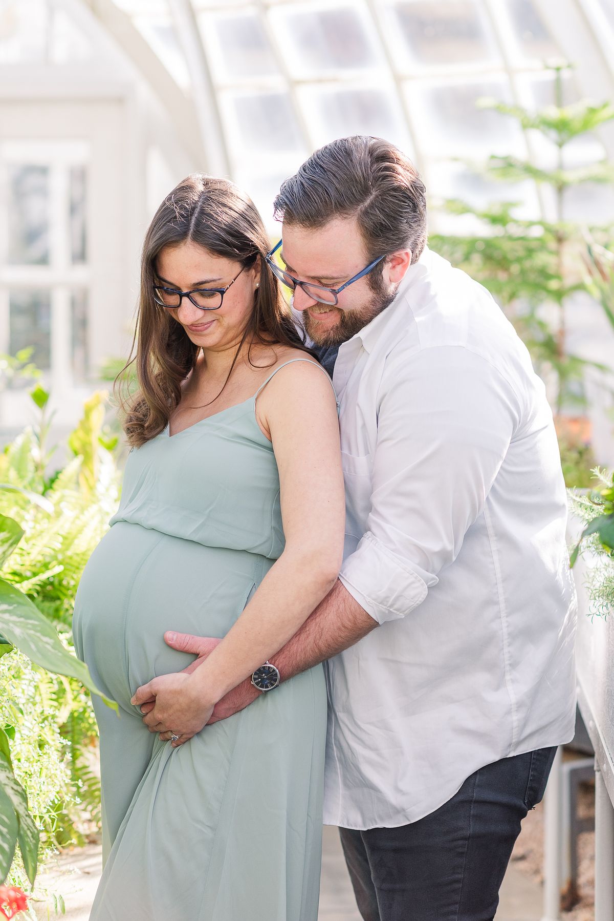 Husband and wife holding her pregnant belly and smiling in The Frick greenhouse