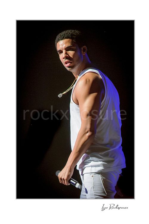 Vertical color image of Drake performing live on stage in a white tank top and white jeans with a handheld microphone and dark concert background