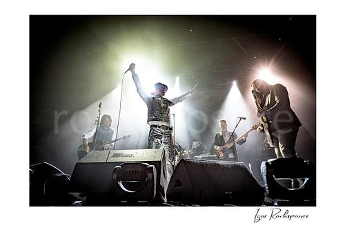 Horizontal color image of The Tragically Hip performing live on stage, with Gord Downie in a glittering silver suit at center with arms raised high while band members play under bright concert spotlights
