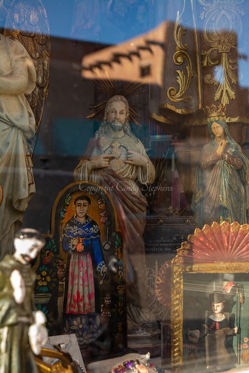 Classic Adobe-style architecture in the background with Catholic religious figures in the foreground, representing Southwestern community traditions.