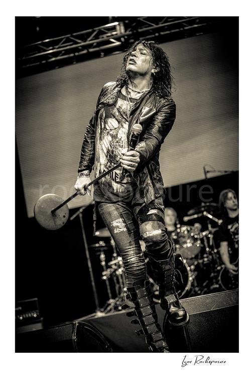 Vertical black and white image of Tom Keifer striding on stage in a dramatic live performance pose, holding a microphone stand with the drum kit visible behind him