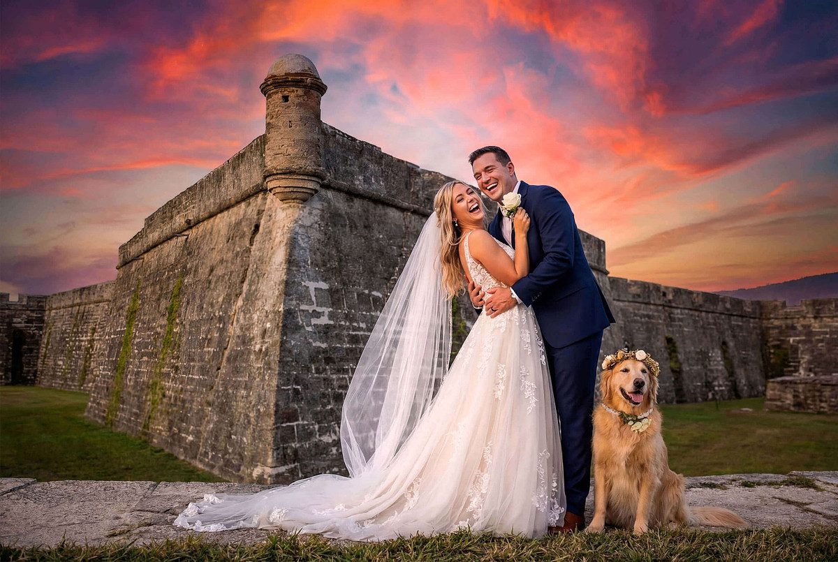 A joyful bride and groom laughing during their sunset elopement at Castillo de San Marcos in St. Augustine, Florida, featuring their Golden Retriever wearing a flower crown.