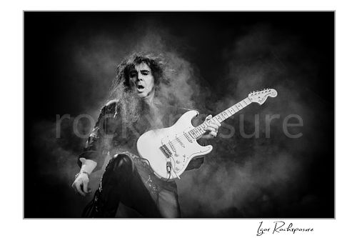 Horizontal black and white image of Yngwie Malmsteen performing live with a white guitar, surrounded by dense stage smoke in a dramatic concert scene