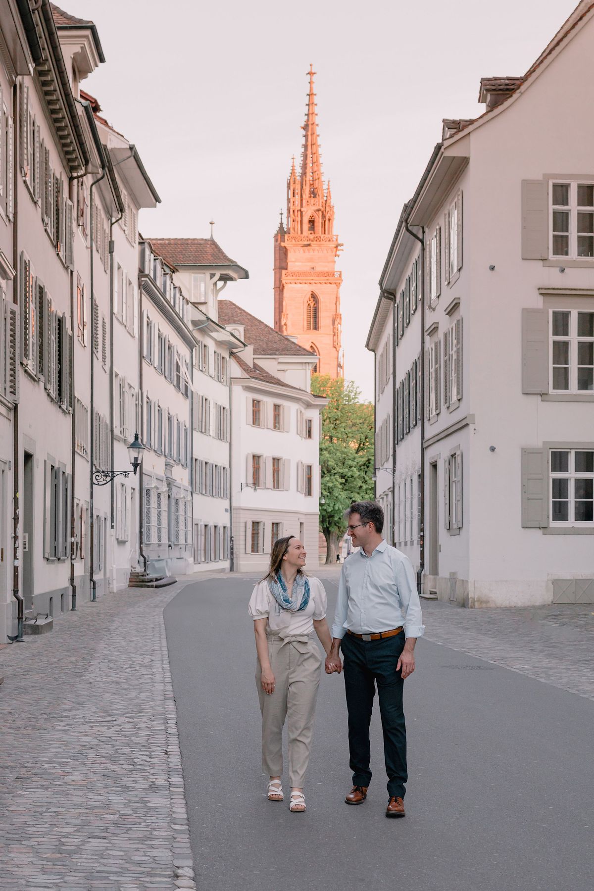 A Basel couple embraces joyfully on a cobblestone street, with a historic Munster cathedral featuring intricate spires in the background. The scene conveys romance and warmth.