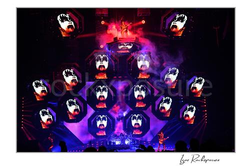 Gene Simmons of KISS standing on an elevated stage platform surrounded by large illuminated Gene Simmons demon face visuals across the stage screens under red and purple concert lighting