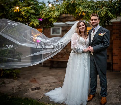 Bride and groom standing side by side, looking at the camera with the bride’s veil flowing in the wind, captured by Weddings By Jermaine.