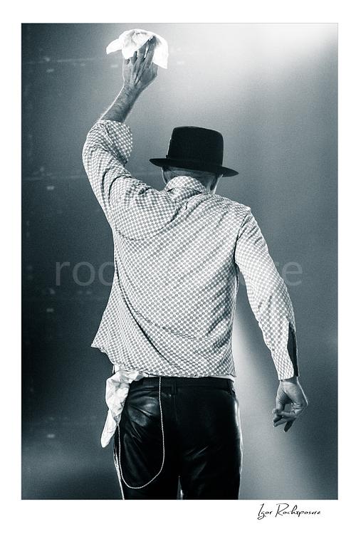 Vertical black and white image of Gord Downie of The Tragically Hip seen from behind on stage, raising a white cloth in a final farewell gesture under soft concert light