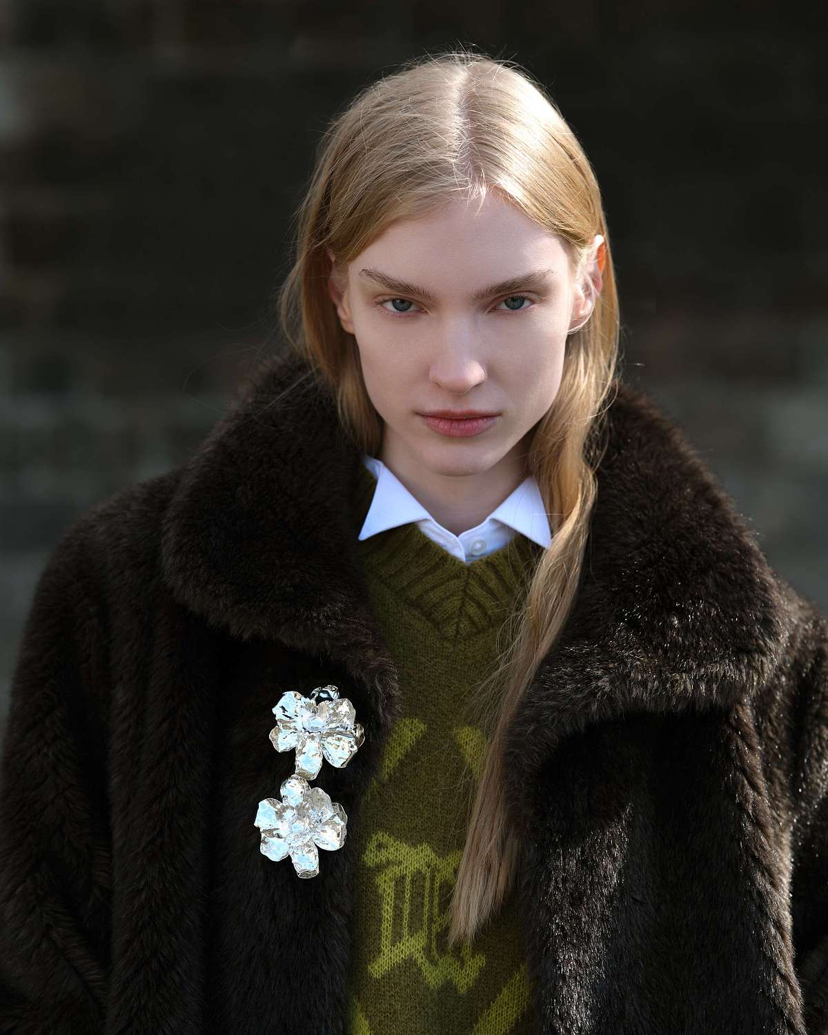 A portrait shot of a woman in a white shirt, lime green sweater, and a fur coat with a brooch, looking straight at the camera