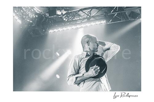 Horizontal black and white image of Gord Downie of The Tragically Hip in an emotional live performance moment, holding his hat to his chest with one hand behind his neck under soft stage light
