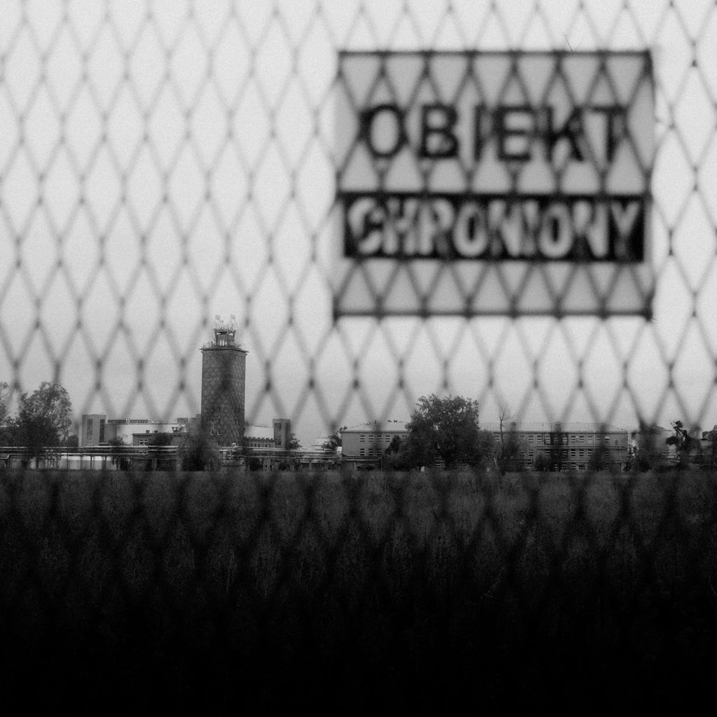 Czarno-biała fotografia przedstawiająca zakład Polfa Tarchomin widziany przez siatkę ogrodzeniową z napisem "Obiekt Chroniony". Zdjęcie wykonane przez Michała Sumkę.