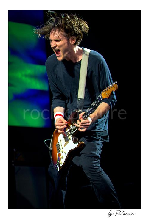 Vertical color image of Josh Klinghoffer of Red Hot Chili Peppers playing guitar in a fierce live performance close-up, with tousled hair and vivid stage color behind him