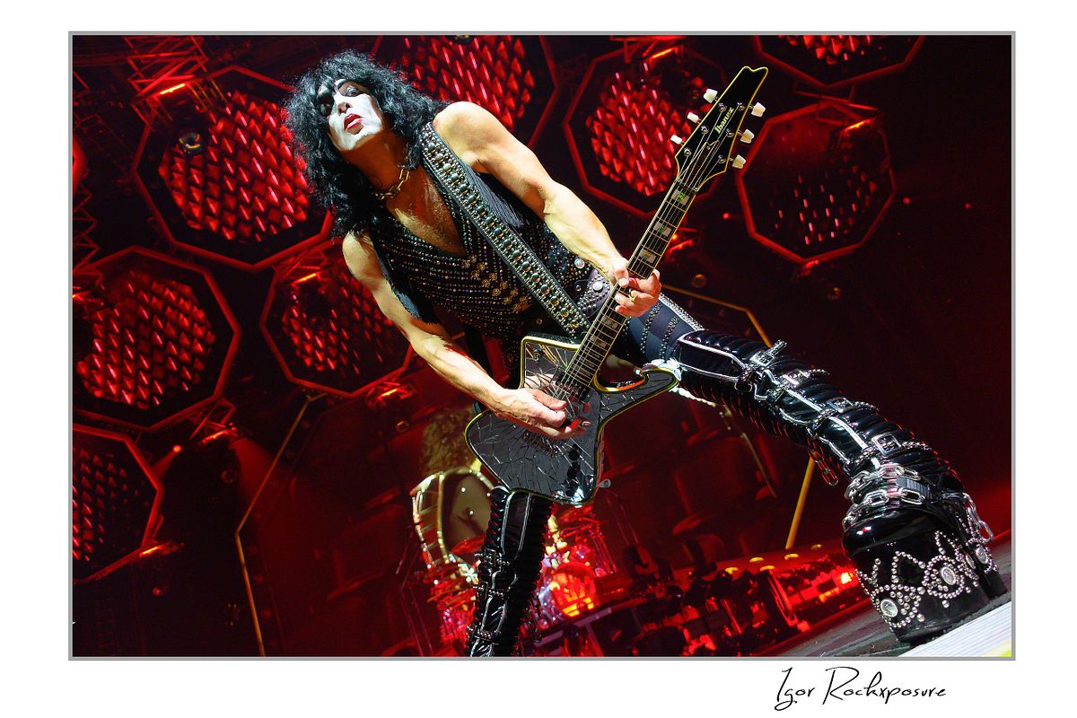 Paul Stanley of KISS performing a wide power stance while playing guitar under intense red stage lighting with dramatic circular stage visuals
