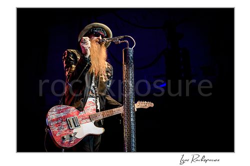 Horizontal color image of Billy Gibbons of ZZ Top performing live on stage with a red Telecaster guitar, singing beside a tall textured microphone stand in his signature hat, dark glasses, and long beard