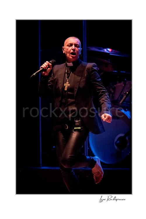 Vertical color image of Sinéad O’Connor singing into a microphone on stage with one foot lifted and the drum kit behind her