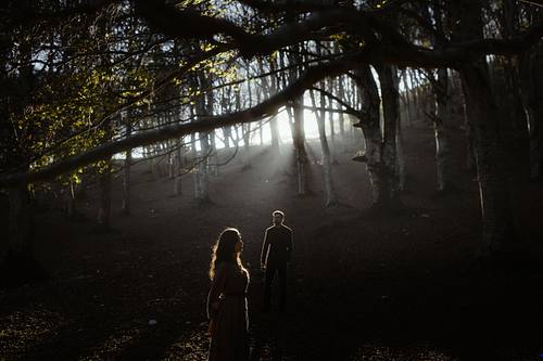 Coppia nel sottobosco marchigiano attraversato da lame di luce, scena ampia e cinematografica.
