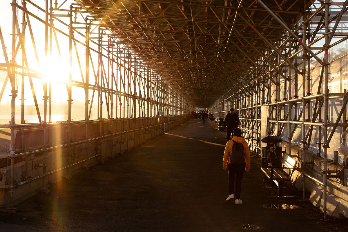L&aring;ng g&aring;ng under byggst&auml;llning i motljus med solstr&aring;lar och m&auml;nniskor som promenerar
