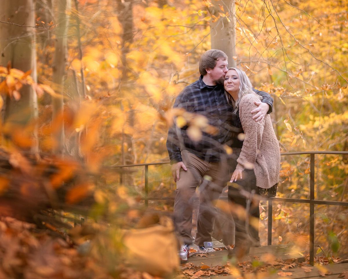 autumn colours in the foreground with the couple sitting on a railing during engagement session