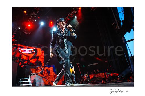 Horizontal color image of Klaus Meine of Scorpions performing live with microphone stand, sunglasses, and vivid red stage lighting