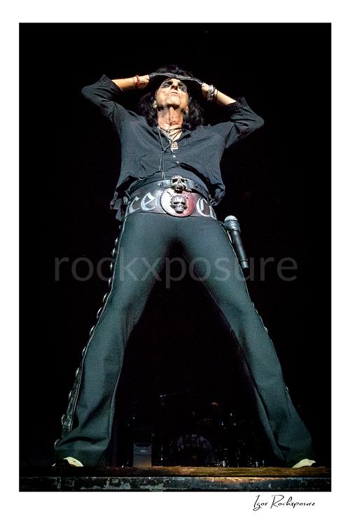 Vertical color image of Alice Cooper performing live on stage in a black outfit with hands raised above his head and a large skull belt buckle