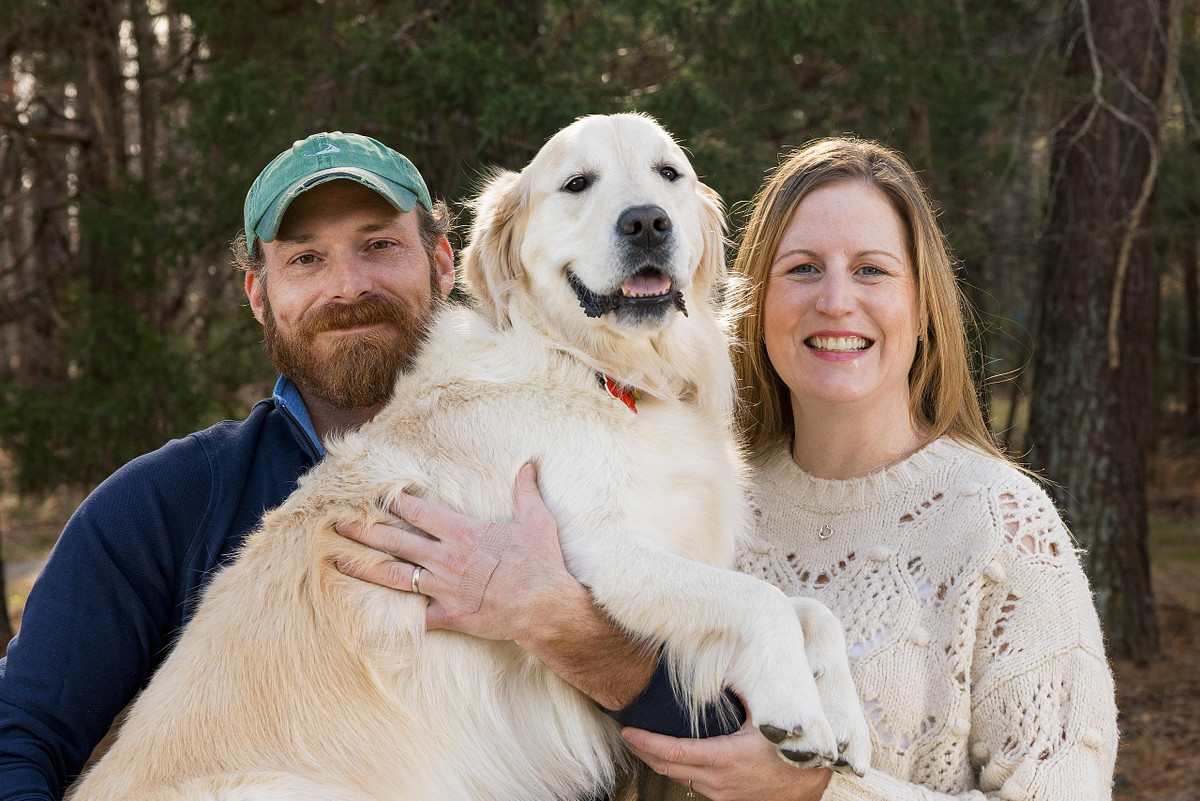 A smiling man in a green hat and a woman in a cream sweater holding their happy Golden Retriever dog during a family photo session in Carrboro, NC.