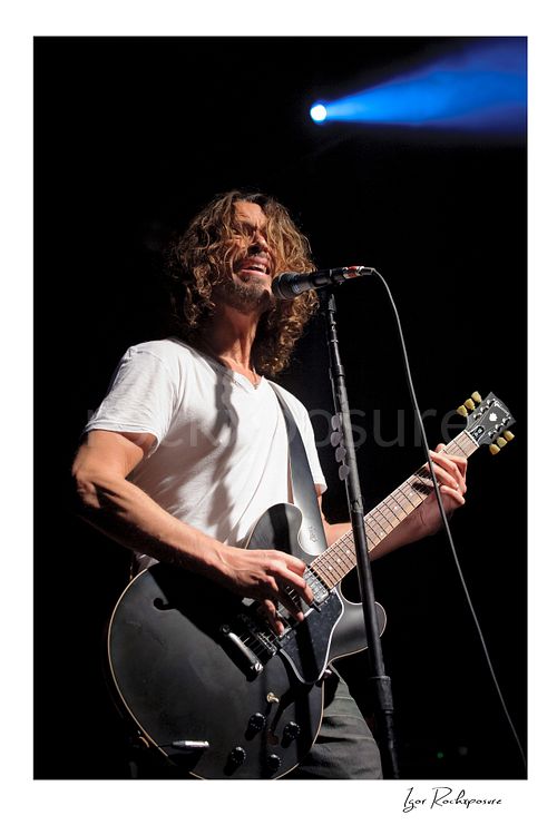 Vertical color image of Chris Cornell performing live on stage, singing into a microphone while playing guitar under blue concert lighting