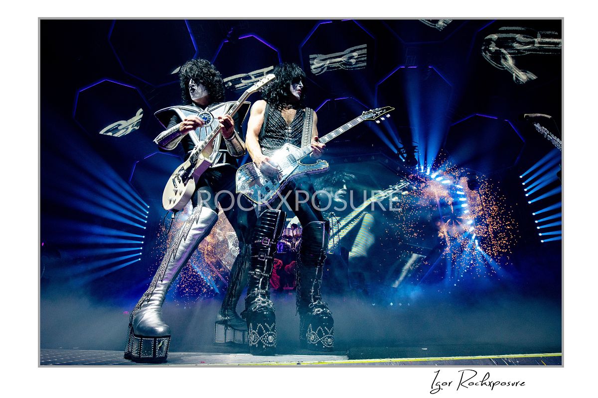Horizontal color photograph of Paul Stanley and Tommy Thayer performing side by side in full makeup and costumes with bright stage lighting, hexagonal screens, and pyrotechnics behind them